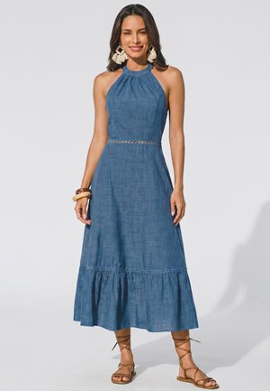 Donna che indossa un abito midi azzurro senza maniche con scollo halter e orlo arricciato, orecchini beige con nappine, braccialetti e sandali marroni con lacci, sorridente.