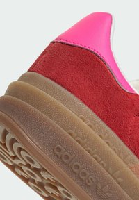 Czerwona zamszowa tenisówka z różowym skórzanym nakładką na pięcie, wyposażona w teksturowaną gumową podeszwę oraz wytłoczone logo „adidas”.