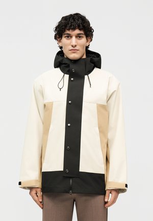 Νέος άνδρας με σγουρά μαλλιά που φοράει μπουφάν σε μπεζ, μαύρες και καφέ αποχρώσεις με κουμπιά μπροστά και κουκούλα.