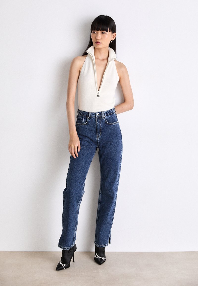 Weißer ärmelloser Body mit hohem Kragen und Frontreißverschluss, kombiniert mit hochtaillierten blauen Jeans und schwarzen spitzen High Heels.