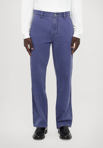 Jean en denim violet avec une coupe droite, design à cinq poches et quincaillerie en métal. Porté avec des chaussures noires et une chemise blanche à manches longues.