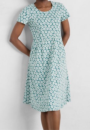 Vrouw draagt een teal-kleurige jurk met korte mouwen tot op de knie met een wit madeliefjesbloemmotief, staand met handen achter de rug tegen een grijze muur.