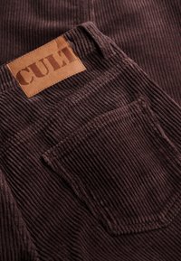 Pantalones de pana marrones con una superficie texturizada, un parche rectangular con la etiqueta "CULT" y un bolsillo trasero.