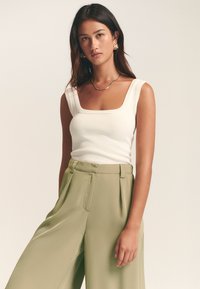 Bela rebrasta majica brez rokavov s štirikotnim izrezom, združena z visokim pasom olivno zelenih hlač z gladko teksturo in stranskimi žepi.