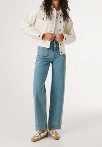 Personne portant un jean large en denim clair, une veste courte blanche, un t-shirt blanc et des baskets beige et blanc, debout devant un fond uni.