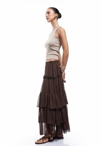 Bruine gelaagde maxi rok met een doorschijnende textuur, vergezeld van een aansluitend beige mouwloos topje. Sandalen maken de outfit af.