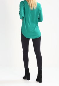 Blusa verde a maniche lunghe con orlo arrotondato, abbinata a jeans skinny neri e stivali alla caviglia. Tessuto morbido con una vestibilità comoda.