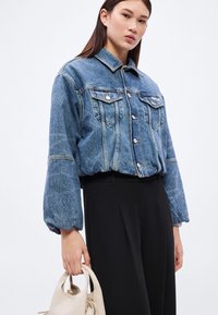 Kék farmer nagy méretű dzseki gombos elejjel, két mellzsebbel és ballon ujjakkal, fekete magasderekú nadrággal és egy világos színű táskával párosítva.