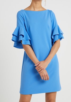 Vrouw gekleed in een helderblauwe jurk met gelaagde ruches aan de mouwen, staand met handen gevouwen voor de taille tegen een effen achtergrond.