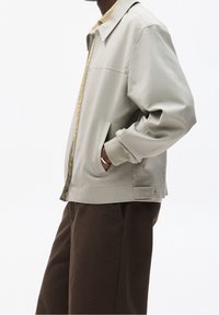 Chaqueta gris clara con cuello puntiagudo, cierre delantero con cremallera y puños acanalados. Cuenta con dos bolsillos laterales y un forro beige en el interior. Combinada con pantalones marrones.