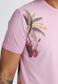 Camiseta de algodón rosa con un diseño impreso de una palmera, un tambor y una guitarra en un lado. Mangas cortas y cuello de tipo crew.