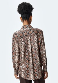 Camicia a stampa pitonata marrone e nera con collettino, maniche lunghe, orlo leggermente curvo e vestibilità rilassata. Tessuto liscio con una texture lucida.