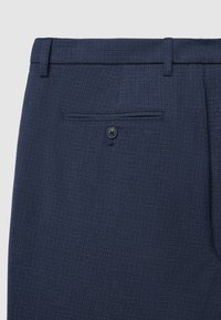 Marineblaue Hose mit feinem Strukturmuster, ausgestattet mit einer Gesäßtasche und einem einzelnen schwarzen Knopf. Maßgeschneidertes Design und eleganter Abschluss.