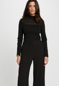 Czarny sweter z wysokim kołnierzem i wzorzystym, koronkowym detalem na ramionach, zestawiony z czarnymi spodniami w szerokim kroju i ozdobnymi mankietami.