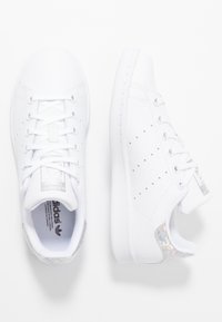 Een paar witte Adidas Stan Smith sneakers met veters, geperforeerde details en holografische hieltabs, weergegeven vanuit boven- en zijaanzicht.