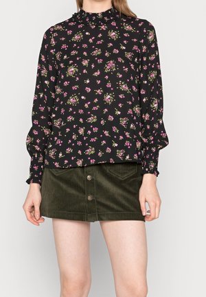 Blusa nera a fiori con fiori rosa e verdi, colletto arricciato e maniche lunghe a sbuffo. Abbinata a una gonna in velluto a coste verde con bottoni.