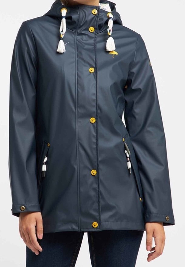 Veste imperméable marine avec capuche, ornée de pompons blancs, de boutons dorés et de deux poches à l'avant. Texture lisse et design moderne.