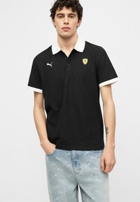 Jeune homme portant un polo noir à col blanc avec les logos Ferrari et Puma, jean bleu clair avec motif Puma, mains dans les poches.