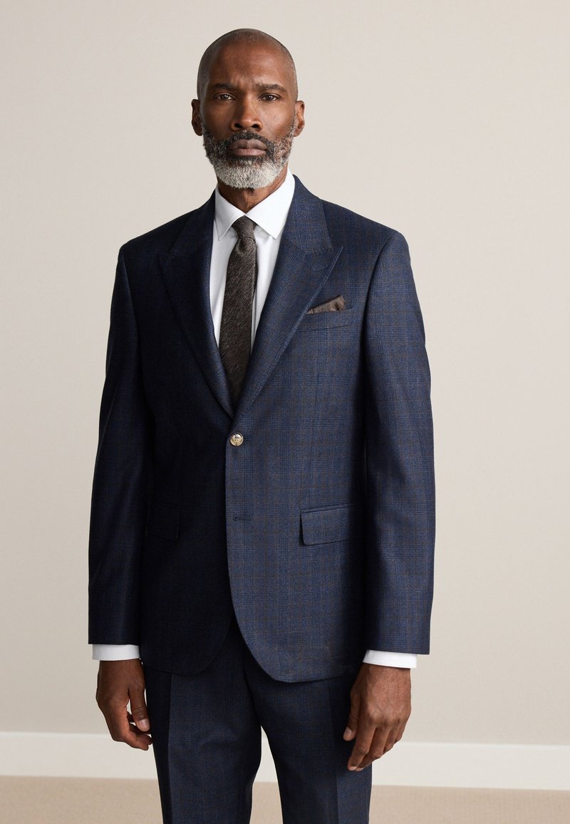 Homme à la barbe grise portant un costume bleu foncé, une chemise blanche, une cravate marron foncé et un mouchoir de poche marron, debout sur un fond beige uni.