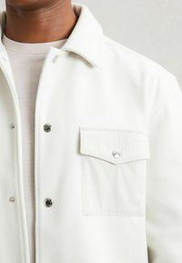 Chaqueta de forro polar blanca con un diseño de cierre de botones, un bolsillo de sarga en el pecho y botones a presión de metal en el frente y el bolsillo.
