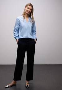 Blouse bleu clair à rayures avec un col en V, coupe décontractée et poignets froncés, associée à un pantalon noir et des chaussures métalliques sans lacets.