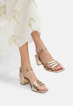 Zlati kovinski sandali na peto imajo štiri široke trakove, trak okoli gležnja z zaponko in kvadratno obliko odprte konice s strukturiranim podplatom.