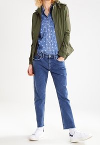 Lekkka oliwkowa kurtka, jasnoniebieska koszula w wzory, niebieskie dżinsy z bocznymi paskami oraz białe trampki z czarnymi akcentami.