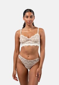 Spitzen-Bralette und String-Set in Crèmefarbe. Mit gewellten Kanten und floralem Muster. Verstellbare Träger und zarte Details überall.