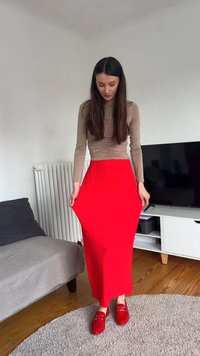 Γυναίκα με μπεζ μακρυμάνικο τοπ, που φοράει φωτεινή κόκκινη φούστα και ταιριαστά κόκκινα παπούτσια, στέκεται σε ένα σαλόνι.