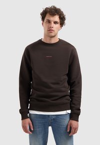 Jonge man met krullend haar, gekleed in een donkerbruin sweatshirt met de tekst "PURE PATH" en een blauwe spijkerbroek, staande tegen een effen achtergrond.