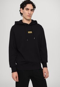 Black hoodie made of cotton blend fabric, featuring a front gold logo patch, drawstring hood, and ribbed cuffs and hem.