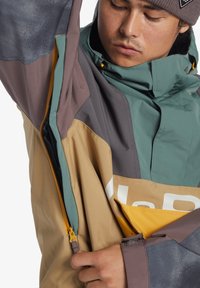Wasserfeste Jacke mit Farbblockierung in grün, braun und grau, einem hohen Kragen und einer seitlichen Tasche mit Reißverschluss und gelbem Ziehband.