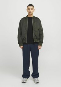 Maslinasto zeleni bomber jakna s rebrastim manžetama, nosi se preko crnog topa. Model nosi široke mornarice hlače i sive sportske cipele. Nema vidljivih uzoraka.