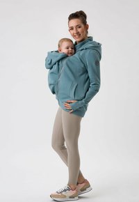 Felpa zip con cappuccio color verde acqua per neonati, dotata di tasca frontale, grande cappuccio, tasche laterali e polsini a coste. Abbinata a leggings beige e sneaker multicolori.