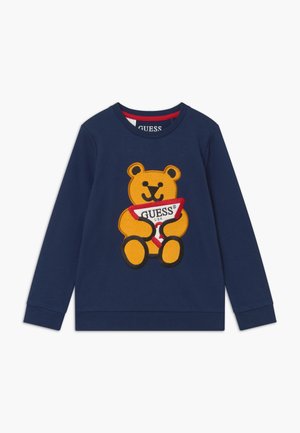 Sudadera azul marino con un gran gráfico de un oso de peluche amarillo sosteniendo una etiqueta triangular roja y blanca con el texto "GUESS". Tejido de mezcla de algodón.