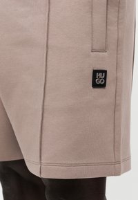 Μπεζ σορτς μέχρι το γόνατο με πλαϊνή τσέπη και μαύρο ορθογώνιο ταμπελάκι με το λογότυπο «HUGO», που φοριέται από ένα άτομο.