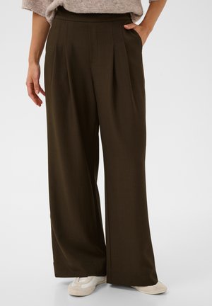 Persoon die een donkerbruine, wijde broek met hoge taille draagt, handen in de zakken en witte sneakers, staand tegen een effen achtergrond.