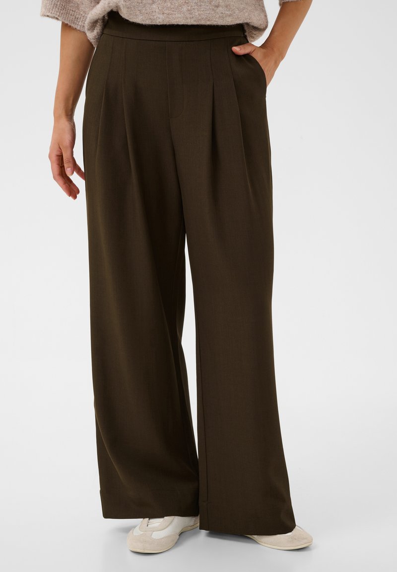 Personne portant un pantalon taille haute, marron foncé, à jambes larges, les mains dans les poches et des baskets blanches, debout devant un fond uni.