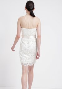 Axelös spetsklänning i off-white med satinmidja; figursydd överdel och vågig fåll. Bakifrån visas ett intrikat spetsmönster.