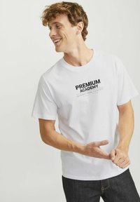 Baltas medvilnės marškinėliai su juodu „PREMIUM ACADEMY“ grafikos spaudiniu, apvaliu iškirpimu, trumpomis rankovėmis ir laisvu siluetu. Paprastas dizainas be jokių papildomų raštų.