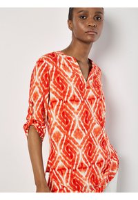 Rote und weiße gemusterte Bluse mit V-Ausschnitt und dreiviertellangen Ärmeln. Verfügt über ein strukturiertes Design und Seitenschlitze für mehr Bewegungsfreiheit.