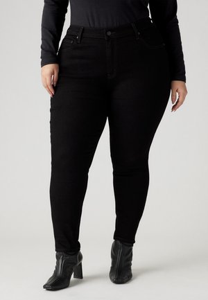 Osoba plus size w czarnych obcisłych dżinsach, czarnej bluzce z długim rękawem i czarnych botkach na obcasie, stojąca na jednolitym tle.