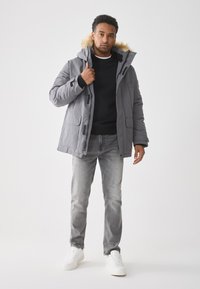 Giacca grigia imbottita con cappuccio foderato in pelliccia sintetica, chiusura con zip, due tasche a pattina, abbinata a un maglione nero, jeans grigi e sneakers bianche.