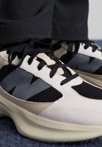 Hvide og sorte sneakers med en tekstureret overdel, sorte snørebånd og en beige sål med et fremtrædende logo på siden.