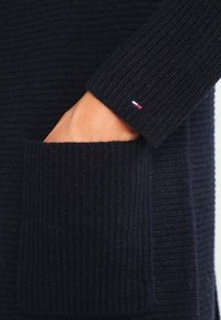 Suéter de punto azul marino con textura acanalada, que presenta un gran bolsillo y un pequeño logo tricolor en el puño. La tela parece suave y cálida.