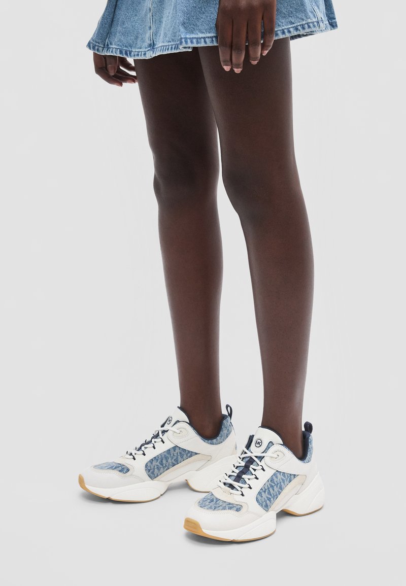 Σκούρα πόδια με λευκά και μπλε sneakers με σχέδια και κοντή τζιν φούστα, που στέκονται σε απλό ανοιχτό φόντο.