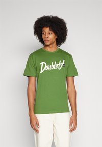 Green cotton t-shirt with white "Doublet" logo, standard fit, round neckline, short sleeves, worn with light-colored pants.