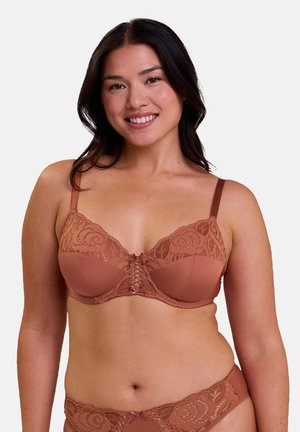 Donna sorridente che indossa reggiseno e mutandine in pizzo color ruggine con motivi floreali abbinati, in piedi su uno sfondo semplice.
