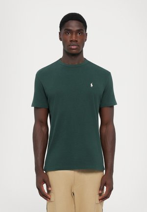 T-shirt en coton vert avec un col rond, des manches courtes et un petit logo brodé blanc sur la poitrine gauche. Texture lisse.