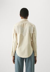 Beige shirt met franjes op de schouders en rug, lange mouwen, kraag en ronde zoom; gedragen met lichtblauwe denim broek.